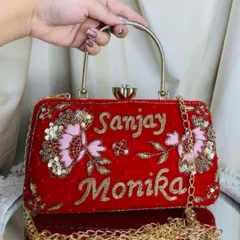 This red velvet clutch features delicate pink and gold floral embroidery alongside the couple's names, 'Sanjay' and 'Monika'. It also comes with a detachable gold chain strap for convenience.