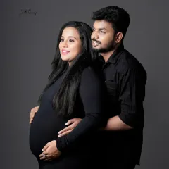 A beautiful and classic couple portrait in matching black outfits. The husband stands behind his wife, gently holding her bump, creating a timeless image of support and love.