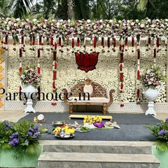 We designed this outdoor wedding stage with a backdrop made entirely of white flowers. The setup is accented with red and white floral hangings, large decorative vases, and lush greenery lining the stage for a fresh, natural look.