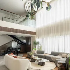 Another view of the viral Kolkata living room, showing the double-height space and the mezzanine level above. The floor-to-ceiling sheer curtains flood the room with natural light, highlighting the custom leaf-inspired chandelier and the grand piano tucked under the stairs.