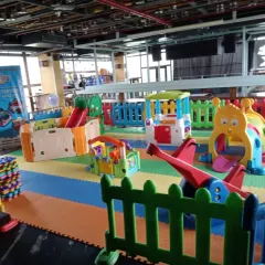 A wide-angle view of an indoor playzone set up in a large hall. The area includes multiple slides, climbers, and soft play structures, all arranged on colorful foam mats to create a safe and engaging environment for kids.