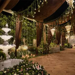 The entrance to a rustic and elegant engagement party. A serene fountain surrounded by flowers and candles leads guests down a path lit by fairy lights, under canopies of brown velvet and hanging greenery.