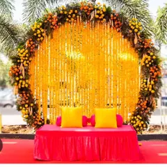 A beautiful outdoor Haldi seating area with a circular backdrop made entirely of marigold strings. The bright yellow flowers against the natural greenery and a simple red-covered seat create a stunning contrast.