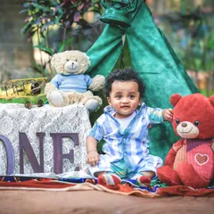 A one-year-old boy sits proudly next to his birthday setup, complete with a "ONE" sign, teddy bears, and a toy plane, ready for his adventure to begin.