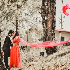 A dynamic and romantic pre-wedding photo in a forest setting. The bride's vibrant red dupatta flows in the wind, creating a beautiful sense of movement as the couple shares an intimate moment.