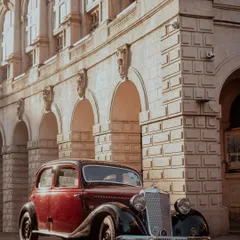 A classic two-tone burgundy and black Mercedes-Benz sedan from a bygone era, parked on a city street. The warm evening light highlights its vintage design against the grand, stone architecture of a heritage building in Mumbai.
