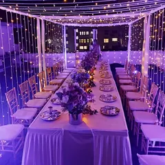The same rooftop dinner setup with purple lighting, showing how we can customize the color scheme to match your event's theme. The table is elegantly set with floral centerpieces and formal place settings.