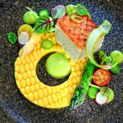 We love playing with textures and shapes in our plating. This dish features a grilled piece of fish served with an artistic corn-kernel ring, asparagus, and fresh greens, creating a visually stunning plate.