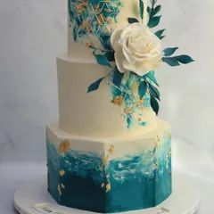 A bold and artistic choice for a modern wedding. This three-tiered cake features a hand-painted watercolor effect in shades of teal, accented with edible gold leaf and a single, striking white sugar rose.