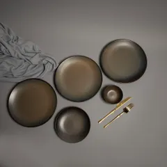 A flat-lay of a rustic, dark-toned ceramic dinnerware set, styled with a grey cloth and gold cutlery for a moody, sophisticated aesthetic.