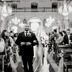The bride walks down the aisle with her brother, her face hidden by the veil in a traditional Catholic custom. This black and white shot captures the anticipation of the moment.
