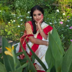 The vibrant red and white of the Bengali saree pop against the lush greenery. This pose, with her hand gently touching her face, adds a touch of classic elegance to the portrait.