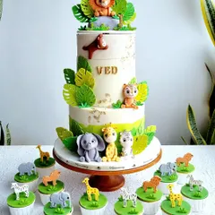 A complete safari-themed dessert setup, featuring a rotating two-tier cake and matching cupcakes. The cake is decorated with cute fondant animals and gold leaf accents, and the cupcakes have individual animal toppers.
