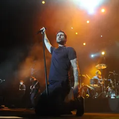 A wide shot of Adam Levine on his knees, completely lost in the music. The warm stage lighting and the band in the background create a complete picture of a live rock performance.