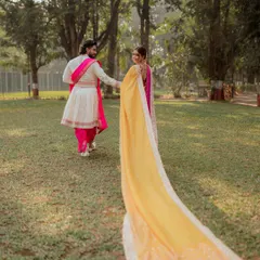 A playful, candid shot capturing the groom leading his bride, her long yellow dupatta trailing behind. This image showcases the beautiful movement and color of traditional Marathi wedding attire in a natural park setting.