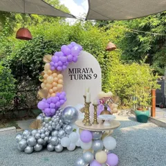 An elegant outdoor birthday setup for "Miraya Turns 9." This decoration includes a personalized arched backdrop, a purple and peach balloon garland, and a table for the cake.