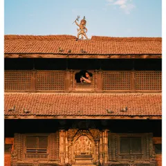 A unique view of a traditional building in Kathmandu, Nepal. The frame captures a deity statue on the roof and a person looking out from a beautifully carved wooden window.