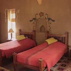 The interior of a twin bedroom in an MP homestay. The space is defined by its earthy mud walls, hand-painted murals above the niche, and locally woven textiles, creating a warm and authentic guest experience.
