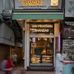 A long-exposure shot of the Shiv Mishthan Bhandar storefront, capturing the motion of the bustling street. The well-lit interior acts as a beacon, drawing customers in from the narrow lane.