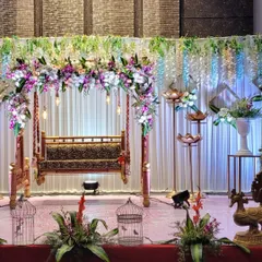 A classic baby shower stage featuring a traditional wooden swing (jhoola) decorated with a beautiful arch of purple and white flowers. The setup is complemented by hanging floral strings and elegant birdcage props on the stage floor.