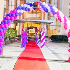 A grand entrance designed for a butterfly themed birthday party. Guests are welcomed through a massive balloon arch in shades of pink, purple, and white, walking down a red carpet towards the main event area.