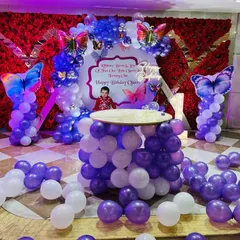 An elaborate butterfly-themed setup in a banquet hall, with a red flower wall, balloon pillars, and a balloon-based cake table.