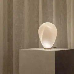 The white glass Solace table lamp emits a soft, diffused glow, its organic form beautifully illuminated against a textured curtain.