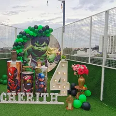 An action-packed Avengers theme party on a rooftop. This setup features a large Hulk backdrop, pedestals with other superhero graphics, a marquee number for the age, and a green and black balloon arch.