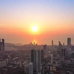 A clear drone shot of the Mumbai cityscape against the Arabian Sea, with the sun setting directly over the water, casting a golden glow.