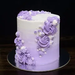 An elegant, tall egg-free cake with a purple ombre finish. The design features a cascade of beautifully piped buttercream roses and swirls in shades of purple and white.