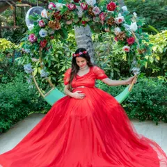 A beautiful moment captured on a garden swing decorated with flowers. The mom-to-be looks serene in a flowing red gown, perfectly complementing the natural green surroundings.