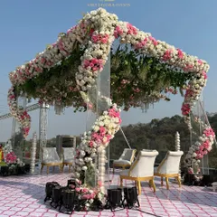 A modern and romantic open air mandap, with spiraling floral arrangements of pink and white roses on transparent pillars. The design allows for an unobstructed view of the natural surroundings.