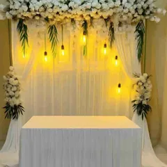 A simple and elegant backdrop for a home-based ceremony or puja, with white drapes, a floral header, and warm hanging bulbs.