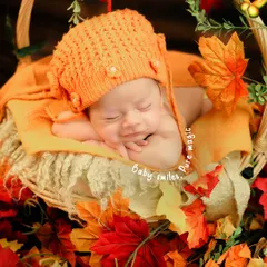 An adorable autumn-themed setup. The baby, wearing a cute orange knitted hat, smiles in their sleep while nestled in a basket filled with colorful autumn leaves.