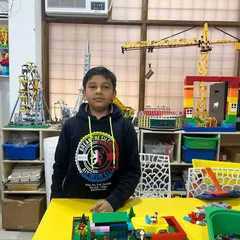A student in our classroom proudly presents his LEGO garden scene. The background is filled with inspiring models, including a large yellow crane and an Eiffel Tower, creating a stimulating learning environment.