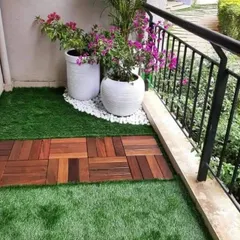 Adding color and life to a small balcony space. This setup features a combination of artificial grass and wooden deck tiles, with flowering bougainvillea and palms in classic white planters.