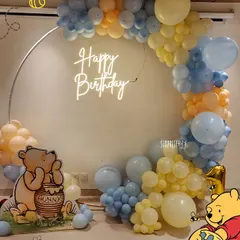 A "pooh-tiful" first birthday setup for little Anaya. This Winnie the Pooh theme featured a round backdrop with a neon "Happy Birthday" sign, surrounded by pastel blue and yellow balloons.