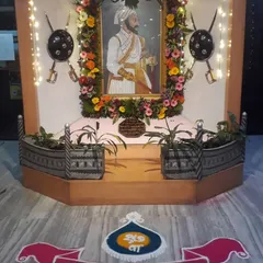 A respectful and beautiful setup for a cultural event, celebrating 'Vardhapan Din'. The decor includes a floral frame for the portrait, traditional shields, and a colorful rangoli.