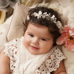 A heart-melting close-up of a smiling baby girl wearing a delicate lace outfit and a floral headband, looking directly at the camera.