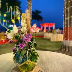 A beautiful table centerpiece for an outdoor evening event. This arrangement includes roses, carnations, and orchids in a geometric glass vase, set on a wooden slice for a rustic yet elegant touch.