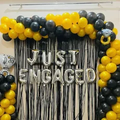 A bold and stylish 'Just Engaged' photo booth. The backdrop features a black tinsel curtain framed by a black and yellow balloon arch, with silver foil balloons announcing the occasion.