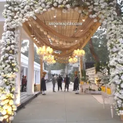 A magnificent floral entrance arch for a daytime celebration. The archway is covered in white flowers, leading into a covered walkway with draped fabric and hanging chandeliers.