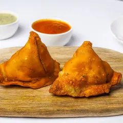 Two perfectly fried samosas on a wooden board, with dipping sauces and onions in the background. This classic Indian snack is shot on a white background, ideal for a clear and appealing menu listing.