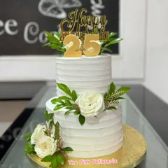 A classic and elegant two-tiered cake for a 25th anniversary. The clean, white textured buttercream is beautifully decorated with fresh white roses, greenery, and a gold "25" topper.