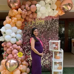 An elegant "Oh Baby" backdrop for a baby shower. This setup features a shimmering sequin wall framed by a cascade of peach, pink, and white balloons, along with transparent "BABY" boxes.