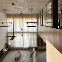 The double-height living room in the Jayanagar Residence creates a sense of grandeur and openness. The mezzanine level with its wood and glass partition maintains a visual connection while defining a separate, more intimate space above.
