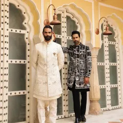 A study in contrasts, this image features two of our signature ceremonial looks. One is a classic off-white embroidered sherwani, and the other is a contemporary black and white patterned bandhgala, showing our range from traditional to modern.