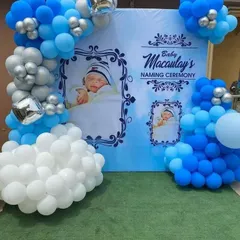 A personalized backdrop for a baby's naming ceremony. This design features a custom printed banner with the baby's photos, framed by a cloud-like arrangement of white, blue, and silver balloons.