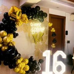 A stylish sweet sixteen birthday setup. The decor features a black and gold balloon ring, a 'Happy Birthday' neon sign, and large light up marquee numbers for the age '16'.