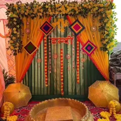 A traditional Haldi or Mehendi backdrop with yellow and green drapes, decorated with marigold flowers and colorful hangings. The setup includes decorative umbrellas and a large brass urli.